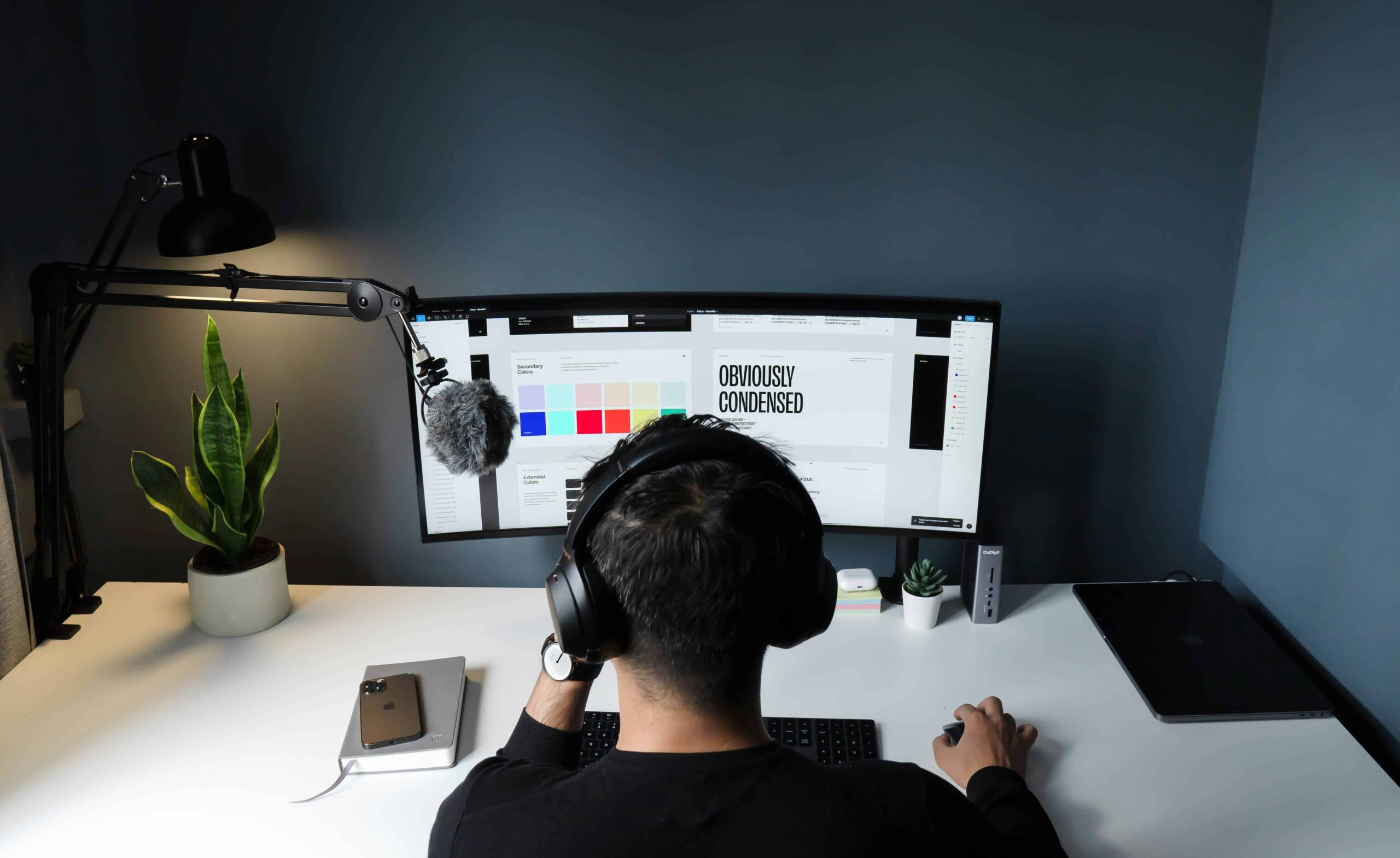 Persoon met koptelefoon van achteren gezien, werkend aan een ontwerp met kleurstalen en typografie op een gebogen monitor.